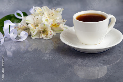 White cup with coffee on a gray background. A bouquet of orchids entwined with a ribbon in the background. Banners, congratulations on the holiday.