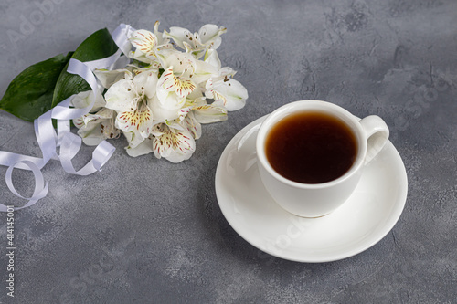 White cup with coffee on a gray background. A bouquet of orchids entwined with a ribbon in the background. Banners, congratulations on the holiday.