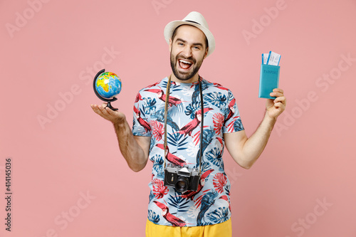 Cheerful young traveler tourist man in summer clothes hat photo camera hold Earth world globe passport tickets isolated on pink background. Passenger traveling on weekend. Air flight journey concept. © ViDi Studio