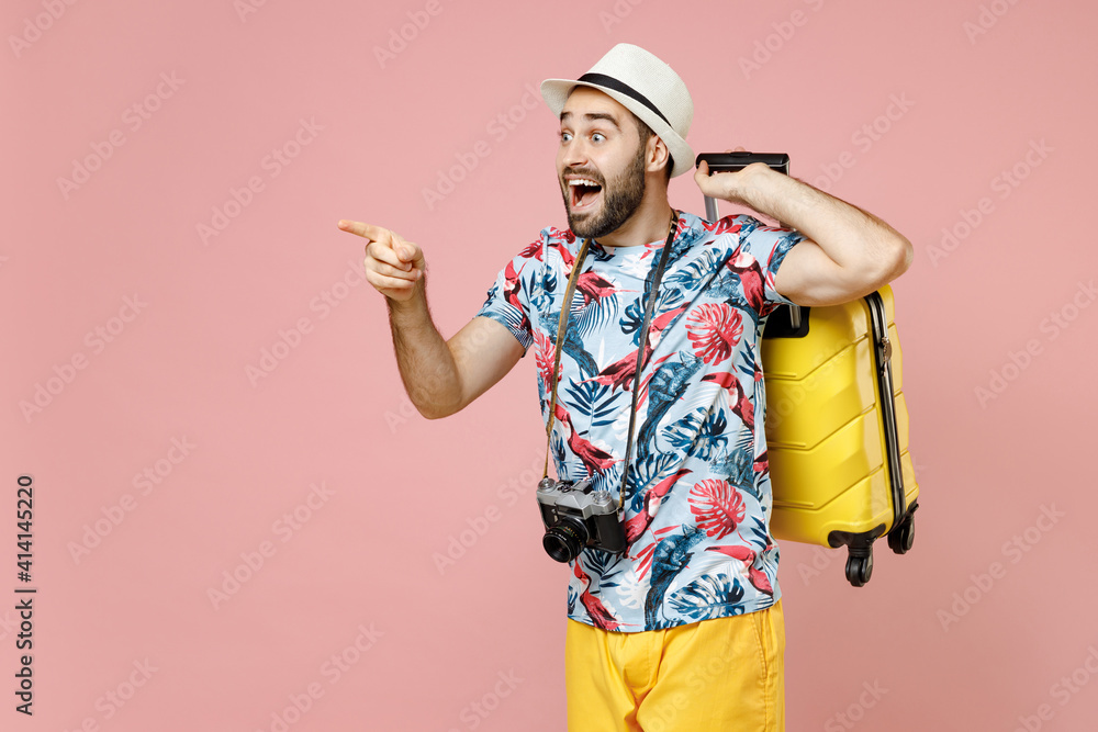 Amazed young traveler tourist man in summer clothes hat photo camera hold suitcase pointing index finger aside isolated on pink background. Passenger traveling on weekend. Air flight journey concept. Amazed young traveler tourist man in summer clothes hat photo camera hold suitcase pointing index finger aside isolated on pink background. Passenger traveling on weekend. Air flight journey concept.