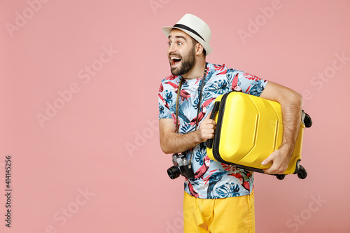 Excited young traveler tourist man in summer basic clothes hat photo camera hold suitcase looking aside isolated on pink background studio. Passenger traveling on weekends. Air flight journey concept.