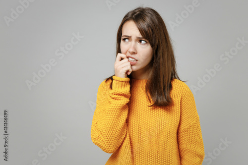 Young thoughtful overthinking pensive attractive troubled woman 20s wearing casual knitted yellow sweater looking aside biting lips nails fingers isolated on grey color background studio portrait photo