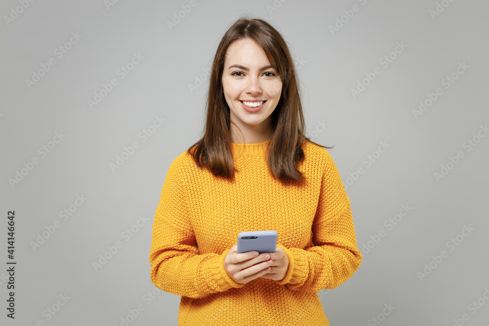 Young smiling cheerful woman 20s wearing casual knitted yellow sweater hold in hand using mobile cell phone chatting browsing typing sms messaging isolated on grey color background studio portrait. Young smiling cheerful woman 20s wearing casual knitted yellow sweater hold in hand using mobile cell phone chatting browsing typing sms messaging isolated on grey color background studio portrait.