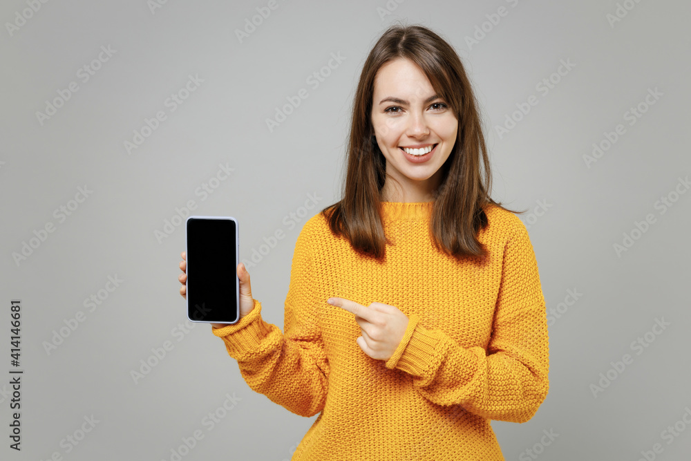 Young smiling attractive beautiful caucasian woman 20s in casual knitted yellow sweater point index finger mobile phone with blank screen workspace area isolated on grey background studio portrait Young smiling attractive beautiful caucasian woman 20s in casual knitted yellow sweater point index finger mobile phone with blank screen workspace area isolated on grey background studio portrait