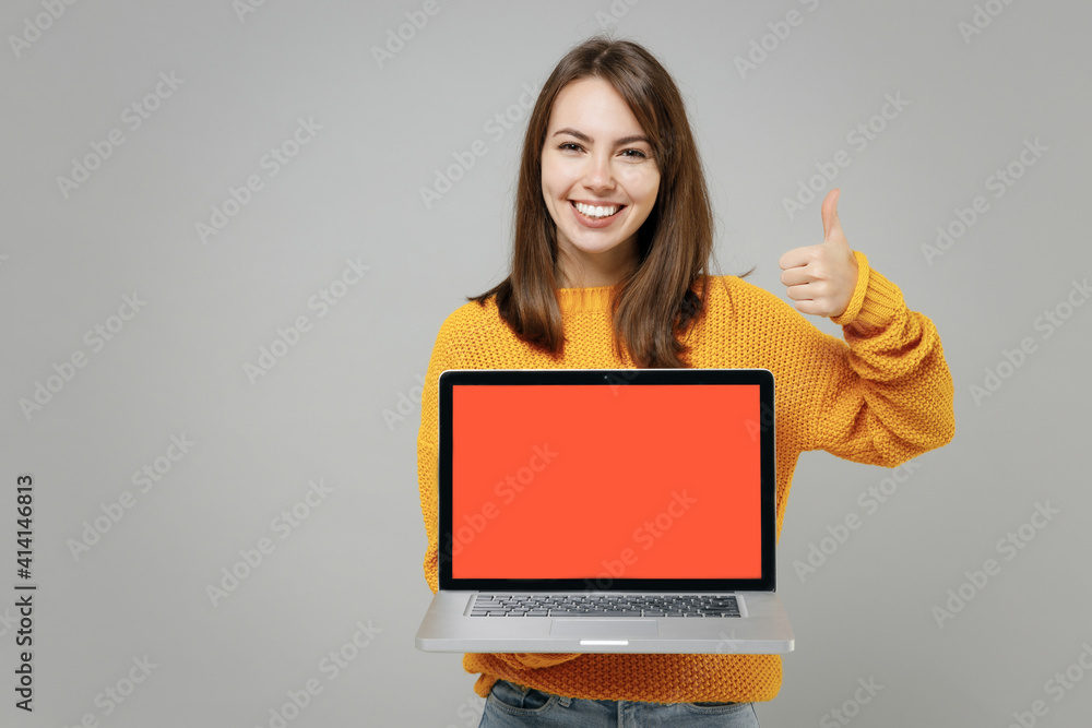 Young smiling freelancer copywriter cute woman 20s in knitted yellow sweater hold laptop pc computer with blank screen workspace area show thumb up gesture isolated on grey background studio portrait Young smiling freelancer copywriter cute woman 20s in knitted yellow sweater hold laptop pc computer with blank screen workspace area show thumb up gesture isolated on grey background studio portrait