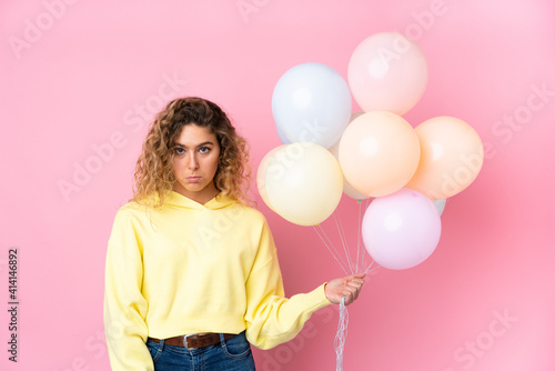 Young blonde woman with curly hair catching many balloons isolated on pink background pleading