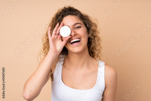 Young blonde woman with curly hair isolated on beige background with moisturizer and happiness