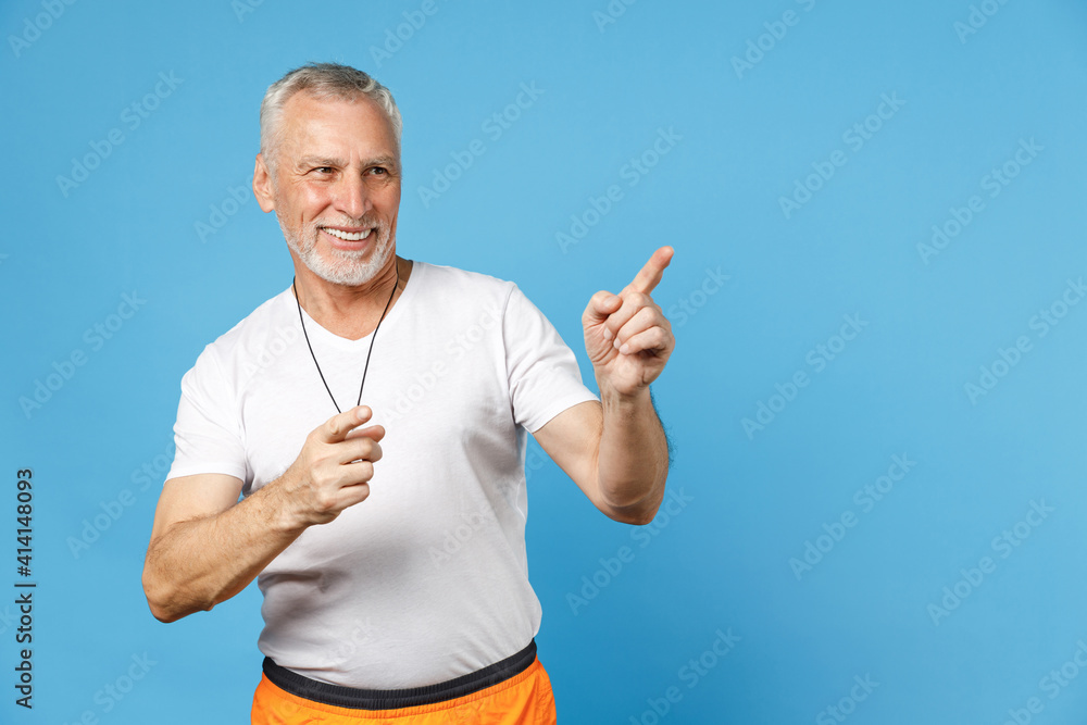 Elderly gray-haired sportsman trainer instructor smiling man 50s in sportswear white t-shirt whistle point index finger aside on copy space isolated on blue background studio. Fitness sport concept Elderly gray-haired sportsman trainer instructor smiling man 50s in sportswear white t-shirt whistle point index finger aside on copy space isolated on blue background studio. Fitness sport concept