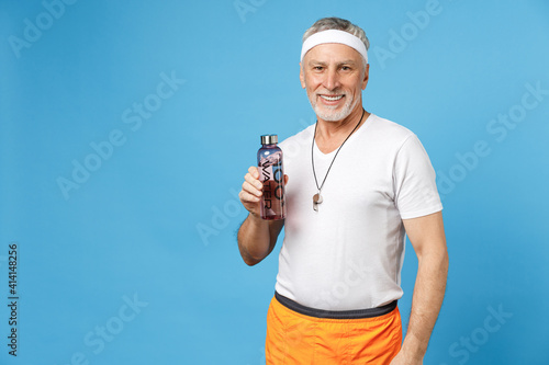 Elderly gray-haired sportsman strong instructor coach smiling man 50s wear sportswear white t-shirt holding water bottle drinking isolated on blue background. Fitness sport lifestyle health concept.