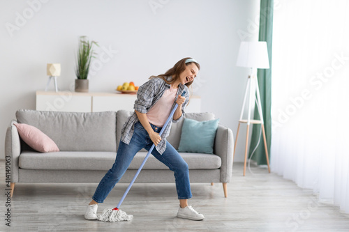 Pretty millennial woman singing while wiping floor at home, using mop as microphone, having fun during domestic duties