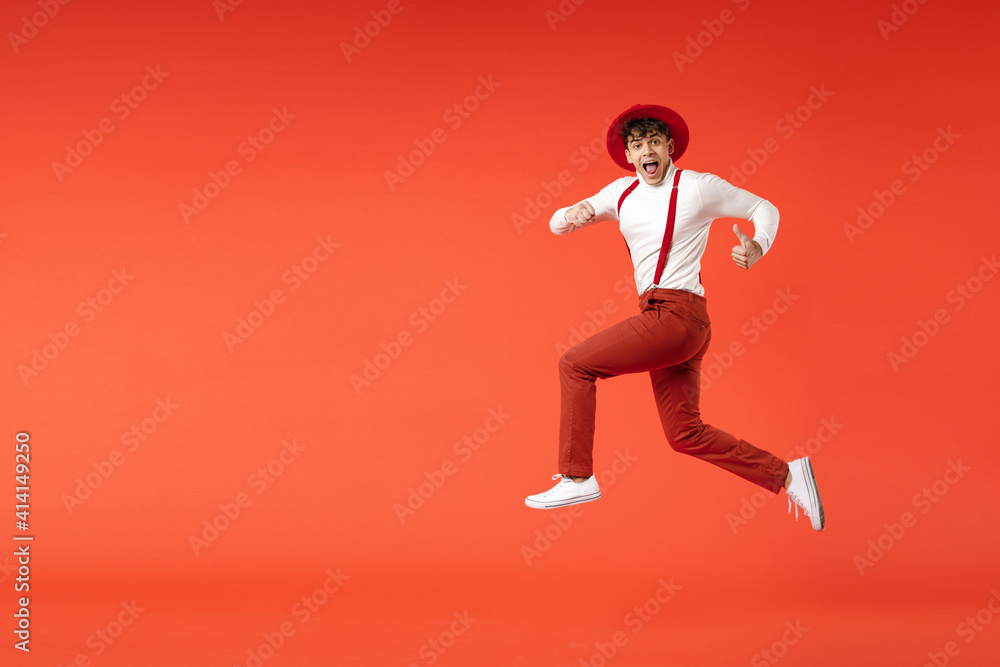 Full length of young spanish latinos smiling cheerful overjoyed fun man 20s wearing hat white shirt trousers, suspenders jump high running fast speed isolated on red color background studio portrait.
