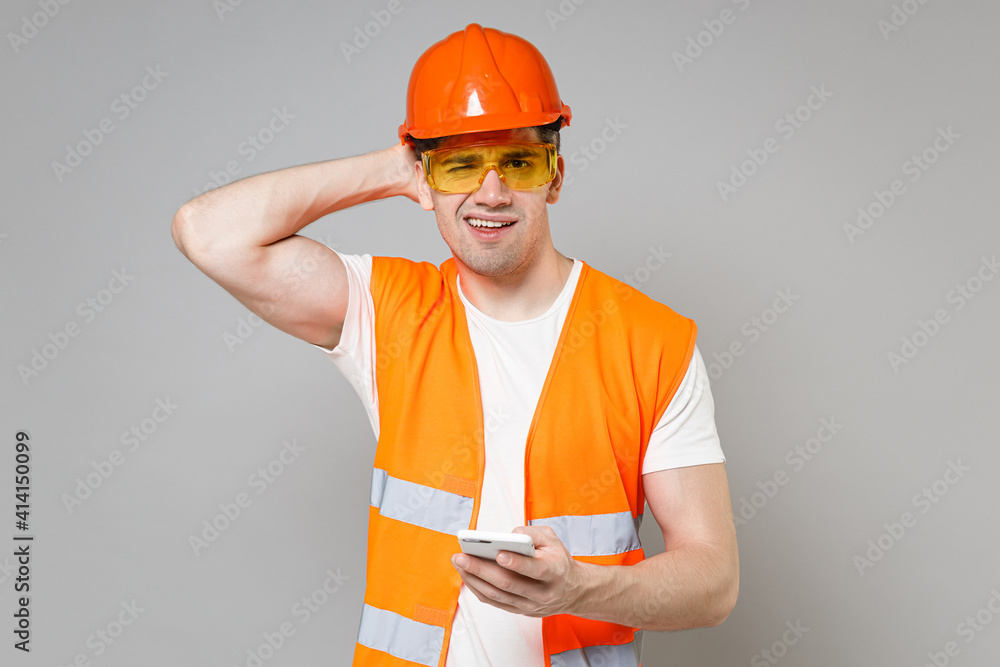 Young employee pensive man in orange vest protective helmet hold mobile phone sctratch head isolated on grey background studio Instruments accessories for renovation apartment Repair home concept Young employee pensive man in orange vest protective helmet hold mobile phone sctratch head isolated on grey background studio Instruments accessories for renovation apartment Repair home concept