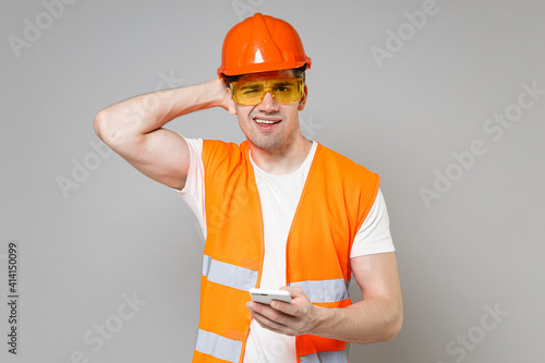 Young employee pensive man in orange vest protective helmet hold mobile phone sctratch head isolated on grey background studio Instruments accessories for renovation apartment Repair home concept photo