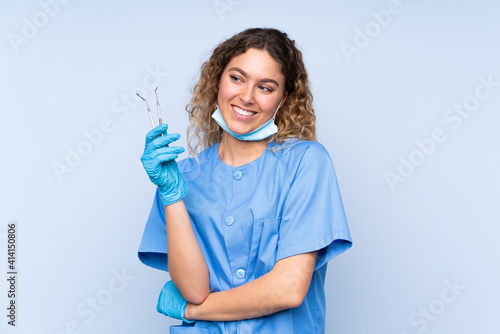 Young blonde woman dentist holding tools isolated on blue background looking to the side