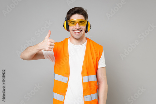 Young employee happy handyman man in orange vest protective ear muffs look camera show thumb up gesture isolated on grey background studio. Instruments for renovation apartment. Repair home concept.