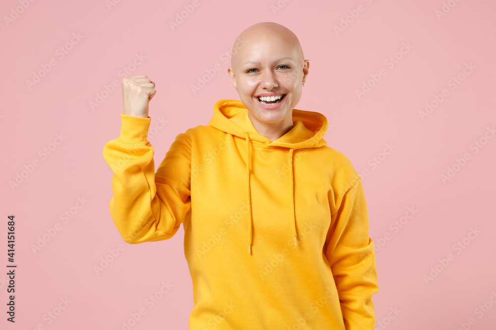 Young bald smiling caucasian happy satisfied woman without hair wearing yellow casual sweatshirt shirt do winner gesture clench fist celebrating isolated on pastel pink background studio portrait. Young bald smiling caucasian happy satisfied woman without hair wearing yellow casual sweatshirt shirt do winner gesture clench fist celebrating isolated on pastel pink background studio portrait.