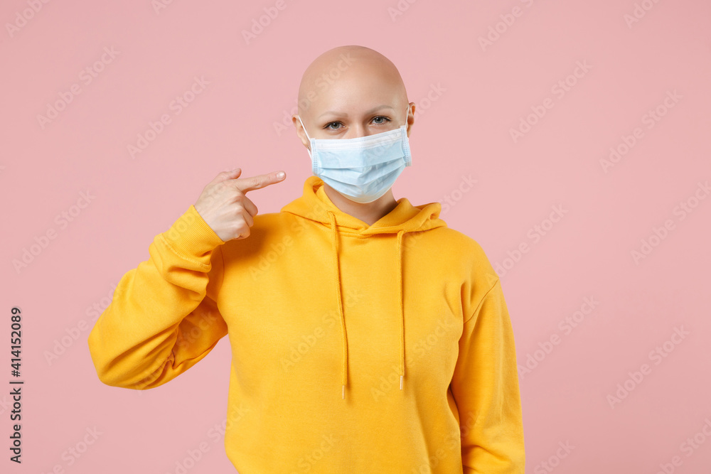 Young bald woman 20s without hair wear yellow casual shirt point index finger on sterile face mask to safe coronavirus virus covid-19, pandemic quarantine isolated on pastel pink background studio