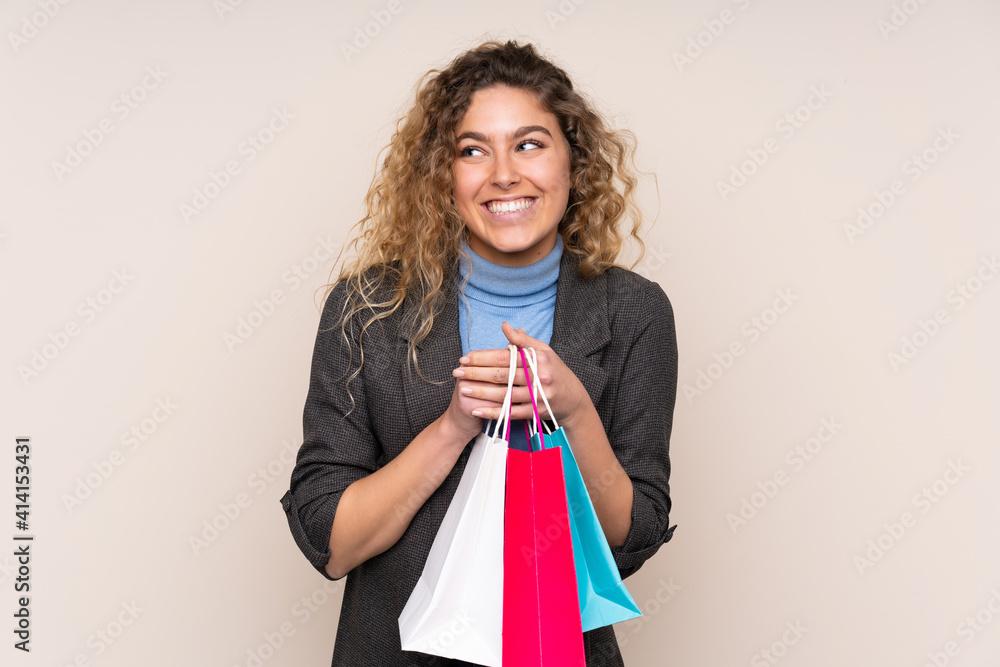 Young blonde woman with curly hair isolated on beige background holding shopping bags and surprised Young blonde woman with curly hair isolated on beige background holding shopping bags and surprised