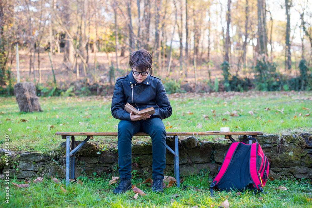 12 year old kid sitting outdoors reads a book, school backpack in the ground-young reader 12 year old kid sitting outdoors reads a book, school backpack in the ground-young reader