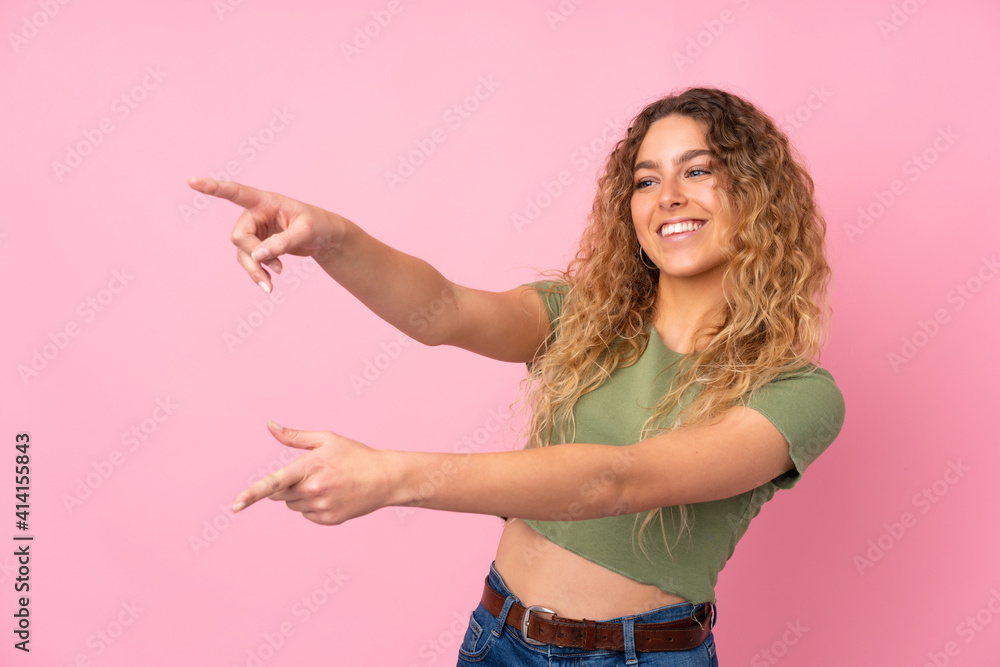 Young blonde woman with curly hair isolated on pink background pointing finger to the side and presenting a product Young blonde woman with curly hair isolated on pink background pointing finger to the side and presenting a product