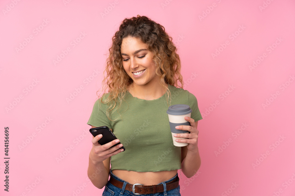 Young blonde woman with curly hair isolated on pink background holding coffee to take away and a mobile Young blonde woman with curly hair isolated on pink background holding coffee to take away and a mobile