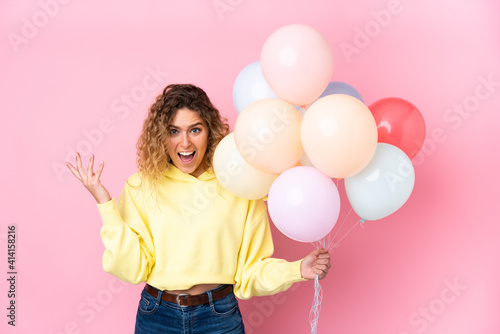Young blonde woman with curly hair catching many balloons isolated on pink background with surprise facial expression