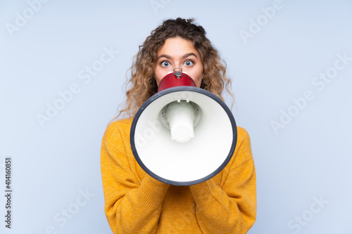 Young blonde woman with curly hair isolated on blue background shouting through a megaphone