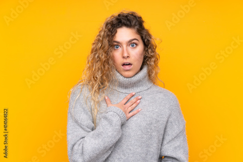 Young blonde woman with curly hair wearing a turtleneck sweater isolated on yellow background surprised and shocked while looking right © luismolinero