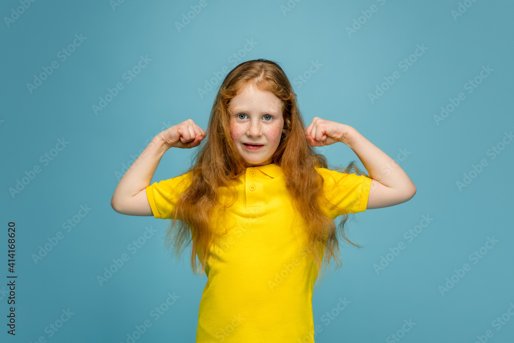 Strong, powerful. Happy, smiley redhair girl isolated on blue studio background with copyspace for ad. Looks happy, cheerful. Childhood, education, human emotions, facial expression concept. Strong, powerful. Happy, smiley redhair girl isolated on blue studio background with copyspace for ad. Looks happy, cheerful. Childhood, education, human emotions, facial expression concept.