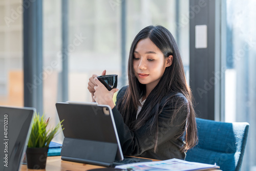 Asian businesswoman sitting at a desk in the office drinking coffee look at a tablet.