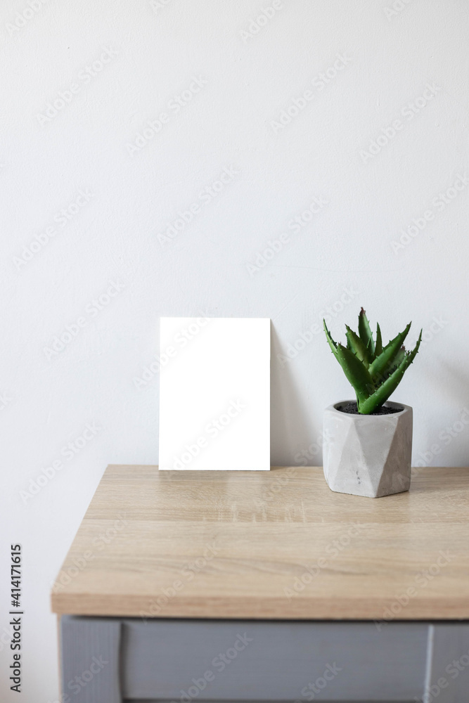 White empty paper blank frame mockup on home wooden table. Modern grey concrete flowerpot with succulent plant. White wall background White empty paper blank frame mockup on home wooden table. Modern grey concrete flowerpot with succulent plant. White wall background