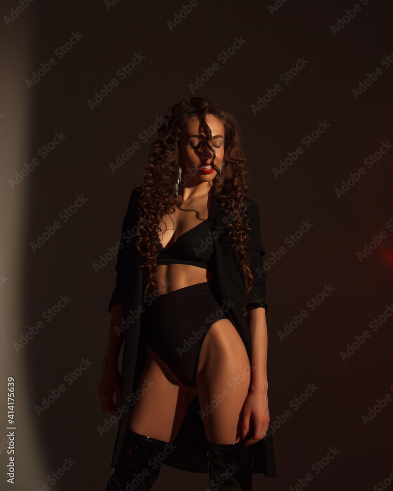 young woman with long curly hair in black lingerie and jacket, in lacquered hessian boots is standing near white wall background with shadows in photo studio, fashion concept, free space young woman with long curly hair in black lingerie and jacket, in lacquered hessian boots is standing near white wall background with shadows in photo studio, fashion concept, free space