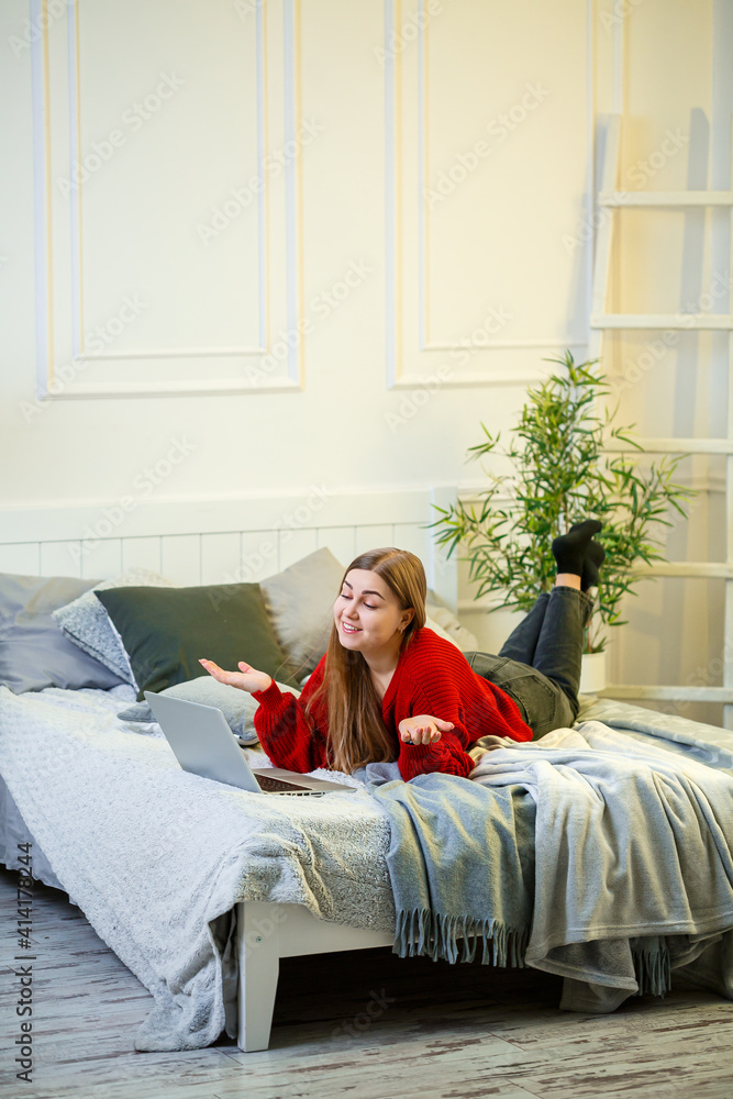 Obraz premium Young woman works at a computer, sitting on a bed, working from a distance. A girl with long hair in a red sweater and jeans works at home.