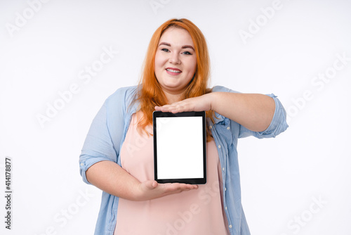 Young plus size red-haired woman showing tablet`s screen isolated over white background photo