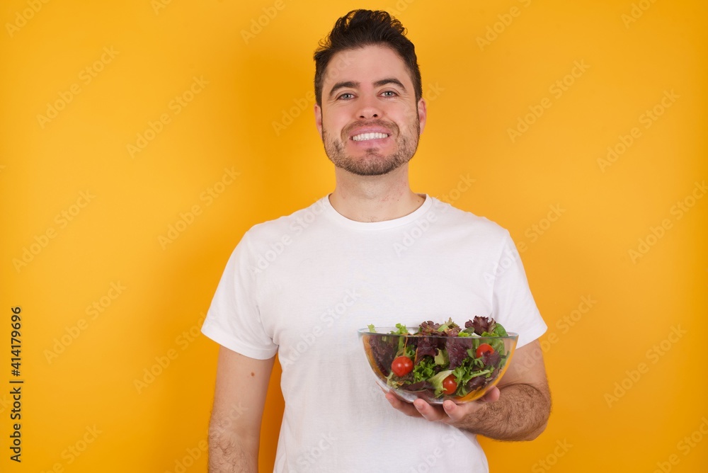 Portrait of successful young handsome Caucasian man holding a salad bowl against yellow wall, smiling broadly with self-assured expression. Confidence and business concept. Portrait of successful young handsome Caucasian man holding a salad bowl against yellow wall, smiling broadly with self-assured expression. Confidence and business concept.