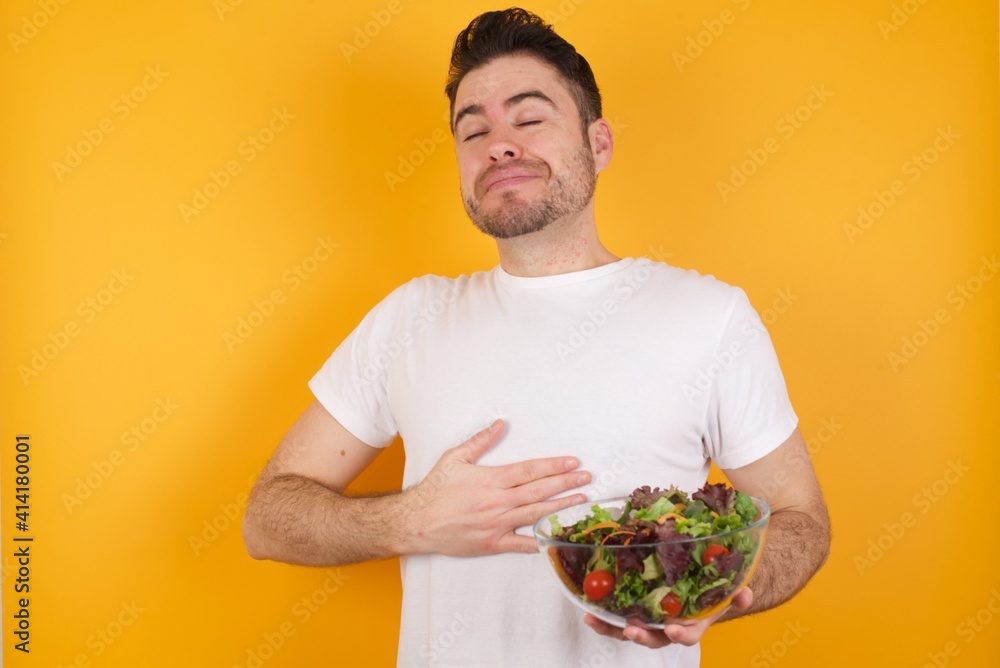 Satisfied smiling young handsome Caucasian man holding a salad bowl, keeps hands on belly, being in good mood after eating delicious supper, demonstrates she is full. Pleasant feeling in stomach. Satisfied smiling young handsome Caucasian man holding a salad bowl, keeps hands on belly, being in good mood after eating delicious supper, demonstrates she is full. Pleasant feeling in stomach.