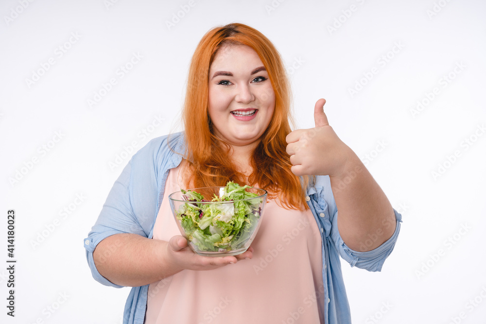 Happy plump woman with red hair showing thumb up with a bowl of salad isolated over white background Happy plump woman with red hair showing thumb up with a bowl of salad isolated over white background