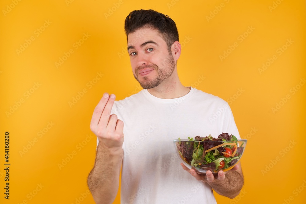 young handsome Caucasian man holding a salad bowl against yellow wall, making money gesture. young handsome Caucasian man holding a salad bowl against yellow wall, making money gesture.