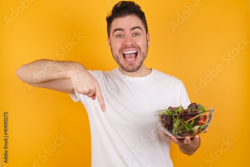 young handsome Caucasian man holding a salad bowl against yellow wall with positive expression, points down with both index fingers, has broad interested smile. Look there, please.
