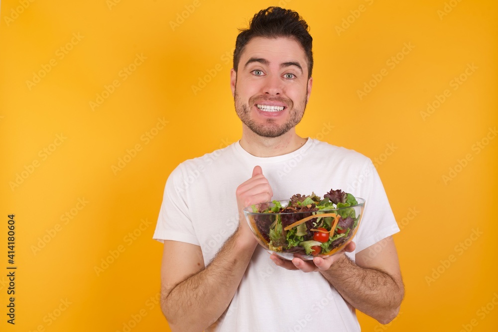 Dreamy charming young handsome Caucasian man holding a salad bowl against yellow wall with pleasant expression, keeps hands crossed near face, excited about something pleasant. Dreamy charming young handsome Caucasian man holding a salad bowl against yellow wall with pleasant expression, keeps hands crossed near face, excited about something pleasant.