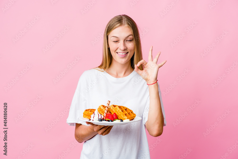 Young russian woman eating a waffle isolated winks an eye and holds an okay gesture with hand. Young russian woman eating a waffle isolated winks an eye and holds an okay gesture with hand.