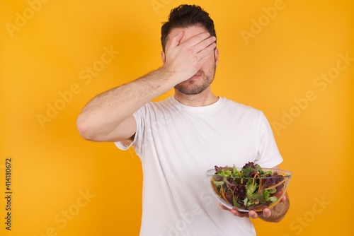 young handsome Caucasian man holding a salad bowl against yellow wall covering eyes with both hands, doesn't want to see anything or feeling ashamed. Human feelings reactions.