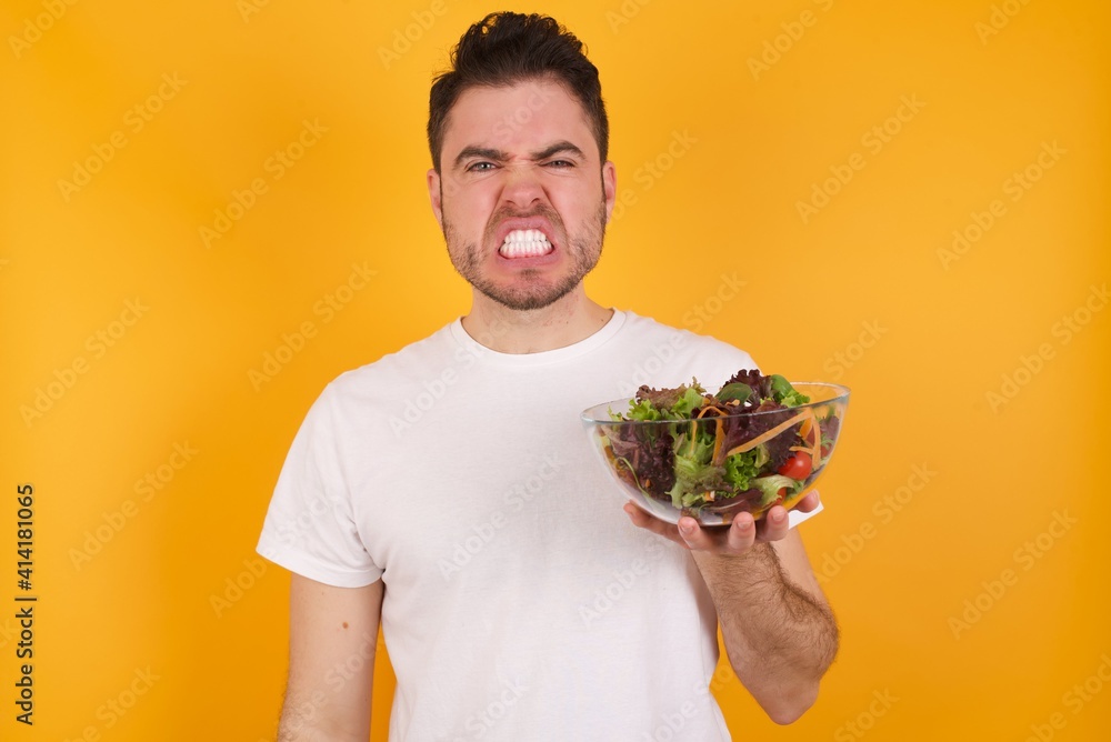 Mad crazy young handsome Caucasian man holding a salad bowl against yellow wall clenches teeth angrily, being annoyed with coming noise. Negative feeling concept. Mad crazy young handsome Caucasian man holding a salad bowl against yellow wall clenches teeth angrily, being annoyed with coming noise. Negative feeling concept.