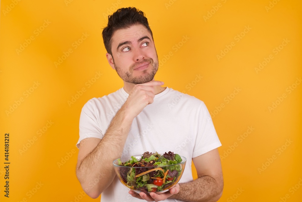 Dreamy young handsome Caucasian man holding a salad bowl against yellow wall with pleasant expression, looks sideways, keeps hand under chin, thinks about something pleasant. Dreamy young handsome Caucasian man holding a salad bowl against yellow wall with pleasant expression, looks sideways, keeps hand under chin, thinks about something pleasant.