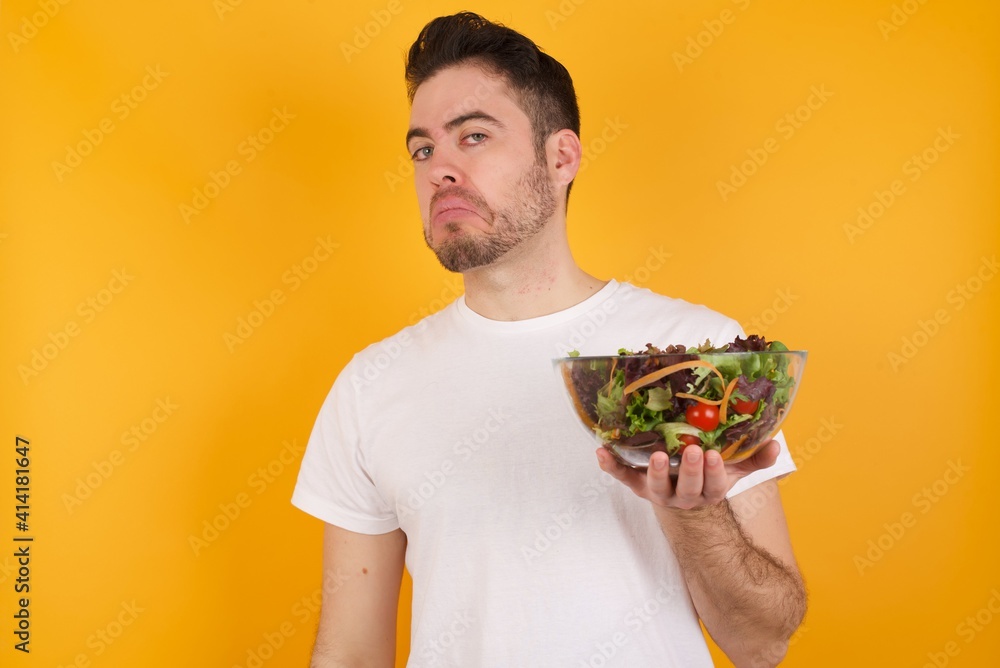 young handsome Caucasian man holding a salad bowl against yellow wall with snobbish expression curving lips and raising eyebrows, looking with doubtful and skeptical expression, suspect and doubt. young handsome Caucasian man holding a salad bowl against yellow wall with snobbish expression curving lips and raising eyebrows, looking with doubtful and skeptical expression, suspect and doubt.