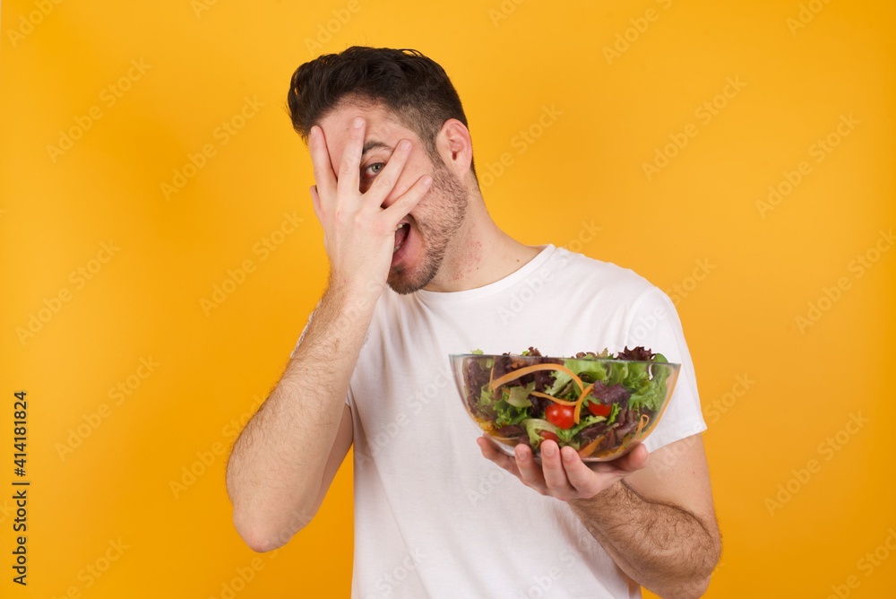 young handsome Caucasian man holding a salad bowl against yellow wall covering face with hands and peering out with one eye between fingers. Scared from something or someone. young handsome Caucasian man holding a salad bowl against yellow wall covering face with hands and peering out with one eye between fingers. Scared from something or someone.