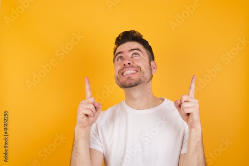 Successful friendly looking young handsome Caucasian man holding a salad bowl against yellow wall exclaiming excitedly, pointing both index fingers up, indicating something.