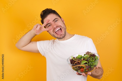 young handsome Caucasian man holding a salad bowl against yellow wall making v-sign near eyes. Leisure, coquettish, celebration, and flirt concept.