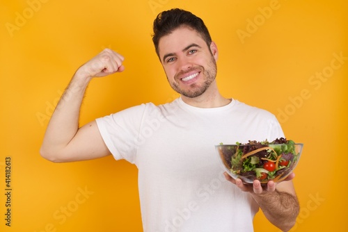 Smiling young handsome Caucasian man holding a salad bowl against yellow wall raises hand to show muscles, feels confident in victory, strong and independent.