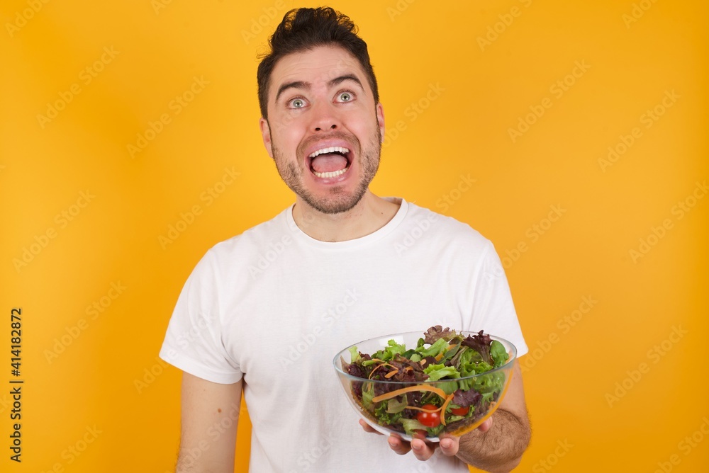 Surprised young handsome Caucasian man holding a salad bowl against yellow wall, shrugs shoulders, looking sideways, being happy and excited. Sudden reactions concept. Surprised young handsome Caucasian man holding a salad bowl against yellow wall, shrugs shoulders, looking sideways, being happy and excited. Sudden reactions concept.